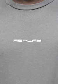 T-shirt cinza feito de algodão macio, com decote redondo. A frente exibe o texto "REPLAY" em fonte branca, limpa e moderna.