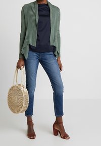 Grön ribbad cardigan, marinblå topp, blå jeans, bruna klacksandaler och en cirkulär vävd väska. Fokusera på textur och lagerdesignselement.