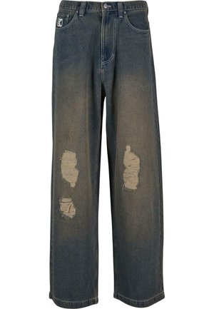 Jeans de denim azul desgastado con pierna ancha, que presentan parches desgastados y bordes deshilachados. Cierre de botón y cremallera de metal, con bolsillos delanteros.