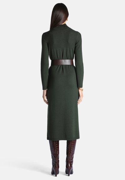 Femme aux longs cheveux foncés portant une robe en maille côtelée vert foncé, une ceinture marron et des bottes en cuir marron remontant jusqu'aux genoux, debout de dos.