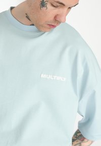 Světle modré oversized tričko vyrobené z měkké látky, s bílým logem "MULTIPLY" na přední straně, s kulatým výstřihem a krátkými rukávy.