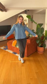 Młoda kobieta w niebieskiej koszuli i czarnych spodniach stoi z rozłożonymi ramionami w jasnym salonie z pomarańczową kanapą i dużą rośliną doniczkową.