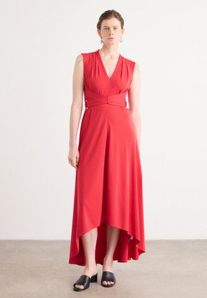 Robe sans manches rouge avec un décolleté en V et un ourlet asymétrique. Le tissu est lisse avec une taille ceinturée mettant en valeur des accents en métal. Sandales noires à glissière.
