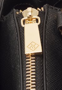 Black fabric zipper with a gold hardware slider and tag, featuring a diamond logo shape. Textured surface detail visible.