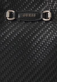 Geantă din piele împletită neagră, cu un logo GUESS proeminent pe o curea texturată, având accente metalice argintii.
