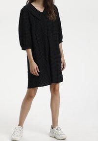 Vestido de cuadros oscuros con un gran cuello, mangas de tres cuartos y un corte holgado. Combinado con zapatillas blancas. Textura de tela suave.