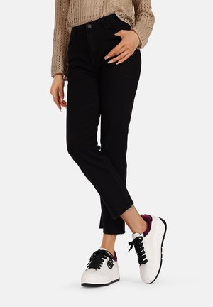 Pantaloni in denim nero con orlo sfrangiato, abbinati a sneakers bianche e nere con un accento viola. Maglione beige lavorato a maglia visibile sopra.