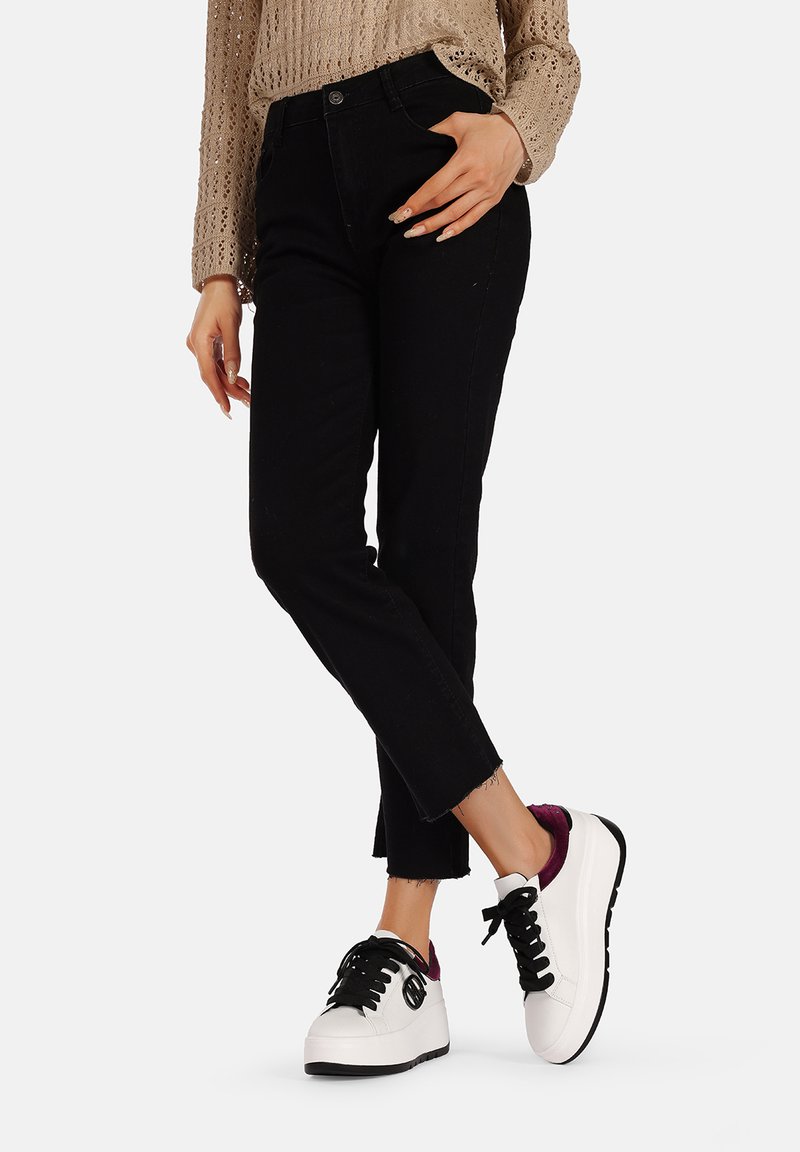 Pantaloni in denim nero con orlo sfrangiato, abbinati a sneakers bianche e nere con un accento viola. Maglione beige lavorato a maglia visibile sopra.