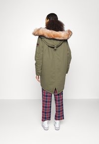 Parka verde oliva con un ampio colletto in pelliccia sintetica, maniche lunghe e uno spacco posteriore. Abbinata a pantaloni a quadri rossi e scarpe da tennis bianche.