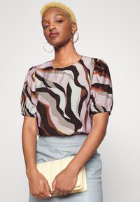 Blusa de manga curta com padrão abstrato em cores pastel e mangas bufantes. A modelo segura uma mala acolchoada amarelo pálido e usa brincos de argola.