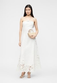 Γυναίκα με λευκό φόρεμα χωρίς τιραντάκια και δαντελένιο τελείωμα, κρατώντας μπεζ τσάντα, στέκεται μπροστά σε απλό φωτεινό φόντο.