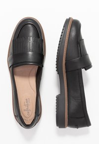 Mocassins en cuir noir avec un design plissé sur le dessus, semelle en caoutchouc marron et profil plat. Doublure douce visible à l'intérieur de la chaussure.