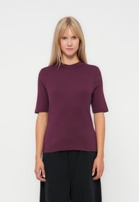 Jeune femme avec des cheveux blonds raides, portant un haut violet à manches courtes et un pantalon large noir, se tenant devant un fond blanc uni.