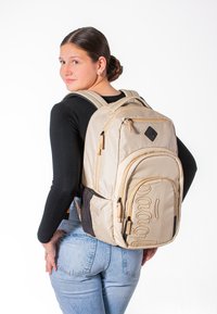Zaino in tessuto beige con grafica frontale, molteplici scomparti con zip, spallacci regolabili e schienale imbottito. Accenti neri sui lati.