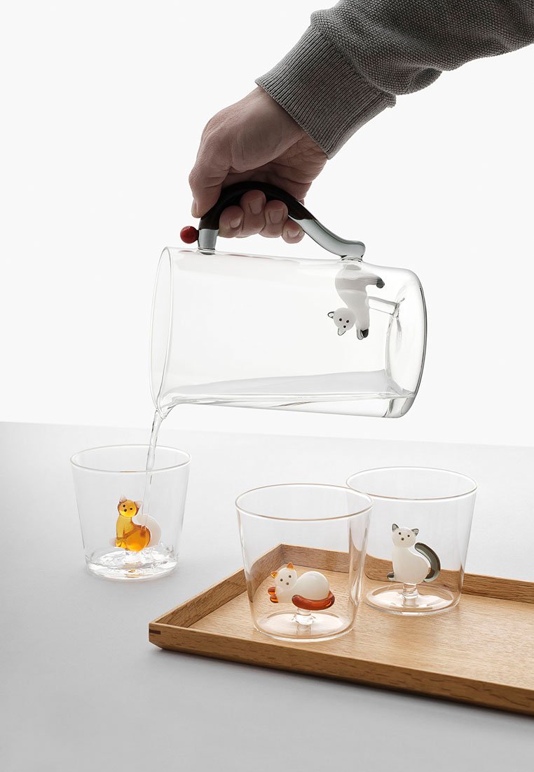 Caraffa in vetro trasparente con manico nero e accenti rossi che versa in tre bicchieri trasparenti con decorazioni di animali. Vassoio di legno sottostante.