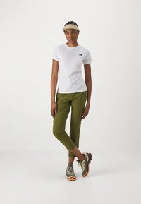 T-shirt bianca a maniche corte con logo, pantaloni color oliva a gamba stretta e scarpe da trekking grigie/verdi con dettagli arancioni, abbinati a una visiera.