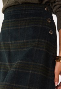 Gonna di panno a scacchi in verde scuro con strisce gialle, caratterizzata da una vita alta e quattro bottoni decorativi lungo il lato. Finitura testurizzata.