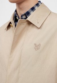 Μπεζ παλτό trench coat με μικρό κεντημένο λογότυπο πουλιού στο στήθος, φορεμένο πάνω από μπλε-άσπρο καρό πουκάμισο.