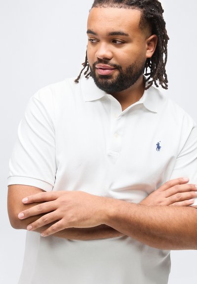 Άνδρας με πλεξουδωτά μαλλιά και γένια, φοράει λευκό πόλο πουκάμισο με σταυρωμένα χέρια, κοιτάζοντας προς τα κάτω και προς τα δεξιά του, σε μονόχρωμο φόντο.