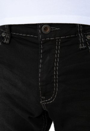 Nærbillede af sorte jeans med hvid kontrastsyning og knaplukning i taljen, båret med en indstukket hvid skjorte.