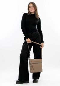 Tan crossbody bag with a smooth texture, featuring a front zip pocket and adjustable strap; worn by a person in black attire.