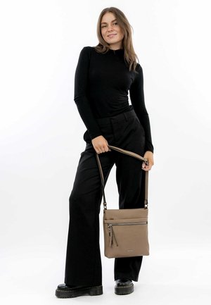Tan crossbody bag with a smooth texture, featuring a front zip pocket and adjustable strap; worn by a person in black attire.