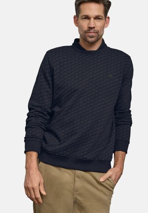 Mand iført en tekstureret marineblå langærmet sweatshirt med ribkant ved ærmerne og khakibukser, stående mod en ensfarvet baggrund.