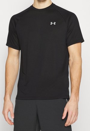Mężczyzna noszący prostą czarną sportową koszulkę Under Armour z krótkim rękawem i czarne spodenki, stojący na jednolitym tle.