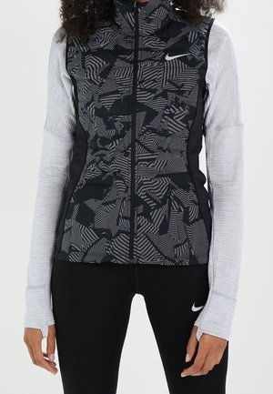 Zwart- en grijsgepatteerd mouwloos vest met ritssluiting over een lichtgrijze top met lange mouwen, gecombineerd met zwarte Nike legging.