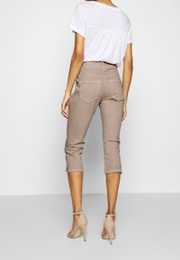 Mujer vestida con pantalones cortos en color topo, blusa blanca de mangas cortas y sandalias de tacón beige, de pie frente a un fondo blanco liso.
