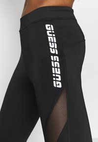 Czarne legginsy z siateczkowym panelem po boku, ozdobione pionowym białym logo "GUESS". Wykonane z elastycznego materiału.