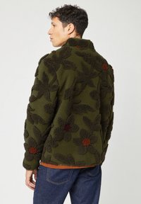 Grön fleecejacka med texturerade blomapplikationer i mörkbrun, orange och vinröd. Hög krage och avslappnad passform. Denimjeans synliga.