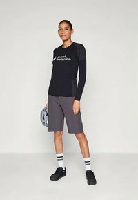 Svart langermet skjorte med "Sweet Protection"-logo, sammen med grå shorts. Svart sko og hvite sokker med grønne striper. Holder en grå hjelm.