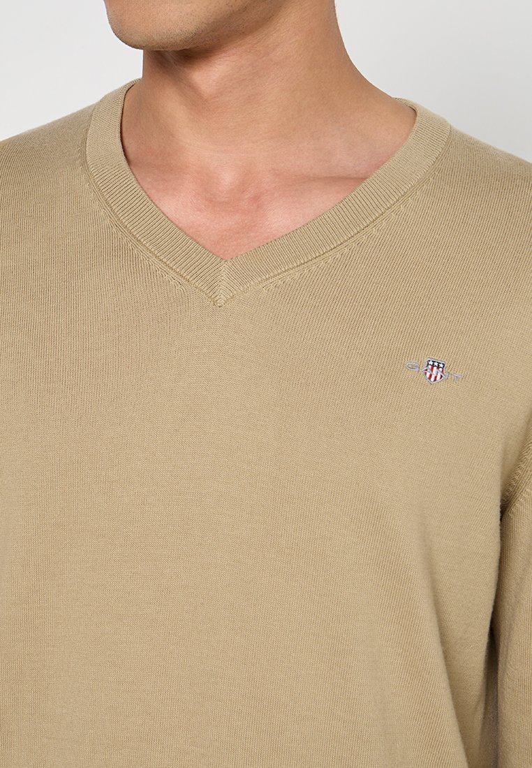 Beżowy sweter z dekoltem w serek, wykonany z dzianiny. Posiada prążkowany kołnierz i mankiety oraz małe, haftowane logo na lewej piersi.