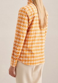 Orange mönstrad skjorta med geometrisk design och vita ovala former. Långa ärmar, knäppningar vid manschetterna och klassisk krage. Slät textur.