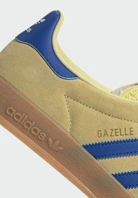 Tenisky Adidas Gazelle v žlutém semišovém materiálu s modrými akcenty a třemi modrými pruhy. Obsahují hnědou gumovou podrážku a detail loga na boku.