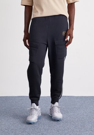 Zwarte cargo joggers met twee voorzakken, elastische tailleband en witte zijkanten, gecombineerd met grijze sportschoenen.