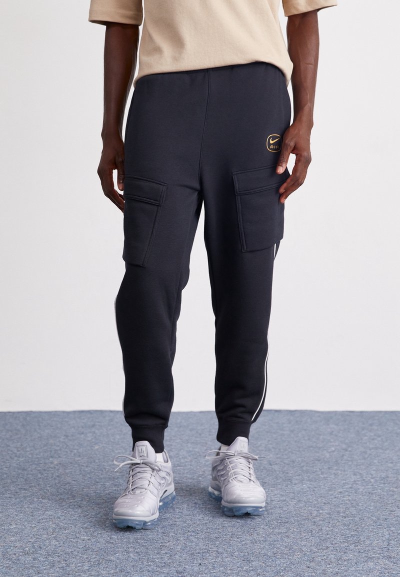 Joggers cargo negros con dos bolsillos frontales, cintura elástica y franjas blancas a los lados, combinados con zapatillas deportivas grises.