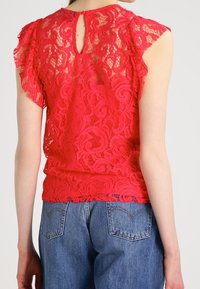 Haut en dentelle rouge avec des manches courtes volantées et un dos en trou de serrure ; associé à un jean bleu en denim. Motif floral complexe sur l'ensemble.
