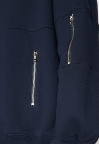 Felpa blu navy con maniche lunghe, dotata di due cerniere laterali e tessuto texturizzato. Il design include polsini a coste e scollo arrotondato.