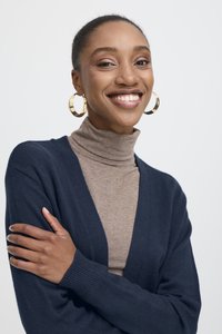 Mosolygó, sötét bőrű nő arany karika fülbevalóval, bézs garbóval és tengerészkék kardigánnal, keresztezett karokkal, egyszínű világos háttér előtt.
