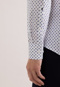 Wit katoenen shirt met lange mouwen en blauwe stippen, voorzien van knoopmanchetten en subtiele geborduurde details op de mouw.