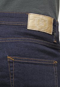 Los vaqueros de mezclilla oscura presentan una etiqueta de cuero marrón en la cintura, resaltada por costuras amarillas a lo largo de los bolsillos y las costuras.