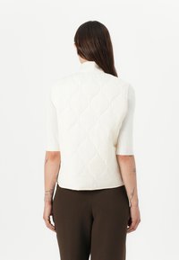 Gilet bianco trapuntato con design aderente, maniche corte e motivo testurizzato, abbinato a pantaloni marroni. Tessuto liscio con un aspetto morbido.