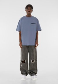 Tricou oversized din bumbac albastru, cu mâneci scurte și un grafic negru pe față, purtat cu blugi gri, largi și uzati, și adidași albi.