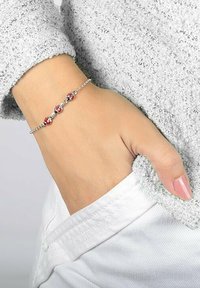 Bracciale in argento con accenti di strass rossi e trasparenti, indossato su una mano vicino a un maglione grigio texturizzato. Un dito poggia su una tasca bianca.