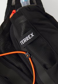 Fekete hálós anyagú felszerelés, amelyen "TERREX" felirat található. Tartalmaz egy vízpalackot, amely narancssárga zsinórral és műanyag klipszel van rögzítve.