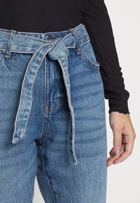 Detailní záběr na modré džíny s vysokým pasem a uvázaným látkovým pásem, které jsou nošeny přes černý žebrovaný top s dlouhým rukávem, ruka uvolněně položená na boku.