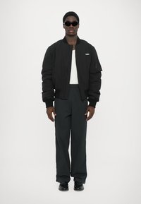 Schwarze Bomberjacke, weißes T-Shirt, dunkelgraue weite Hose und schwarze Schuhe, mit Sonnenbrille. Minimalistisches Design, keine sichtbaren Muster.