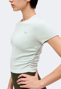 Femme portant un haut Puma à manches courtes ajusté vert clair avec des fronces sur les côtés et des leggings vert foncé, debout avec la main sur la hanche.